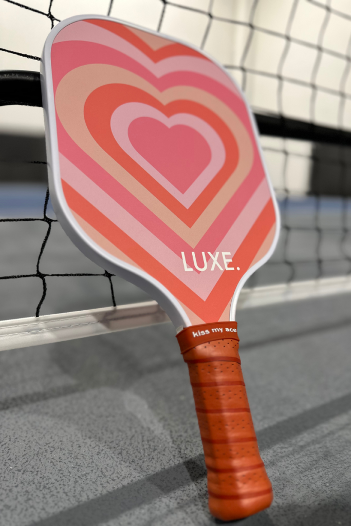 Luxe pickleball paddle review: Pink heart paddle with a brown grip leaning against a pickleball net.