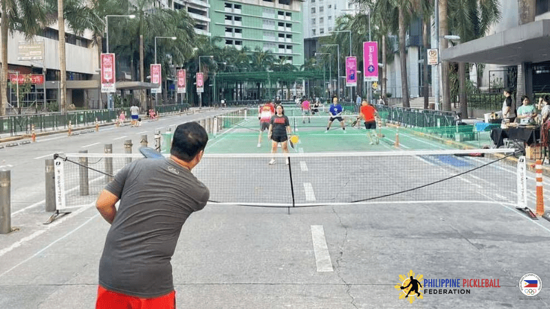 Philippine pickleball federation news: couple playing on a street court on Emerald Avenue in Manila. Man and women with grey shirts, hitting a yellow pickleball over a white and black net.